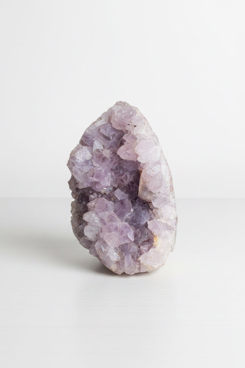 A raw amethyst crystal geode specimen with light purple and pink crystalline formations, standing upright on a white surface against a white background.