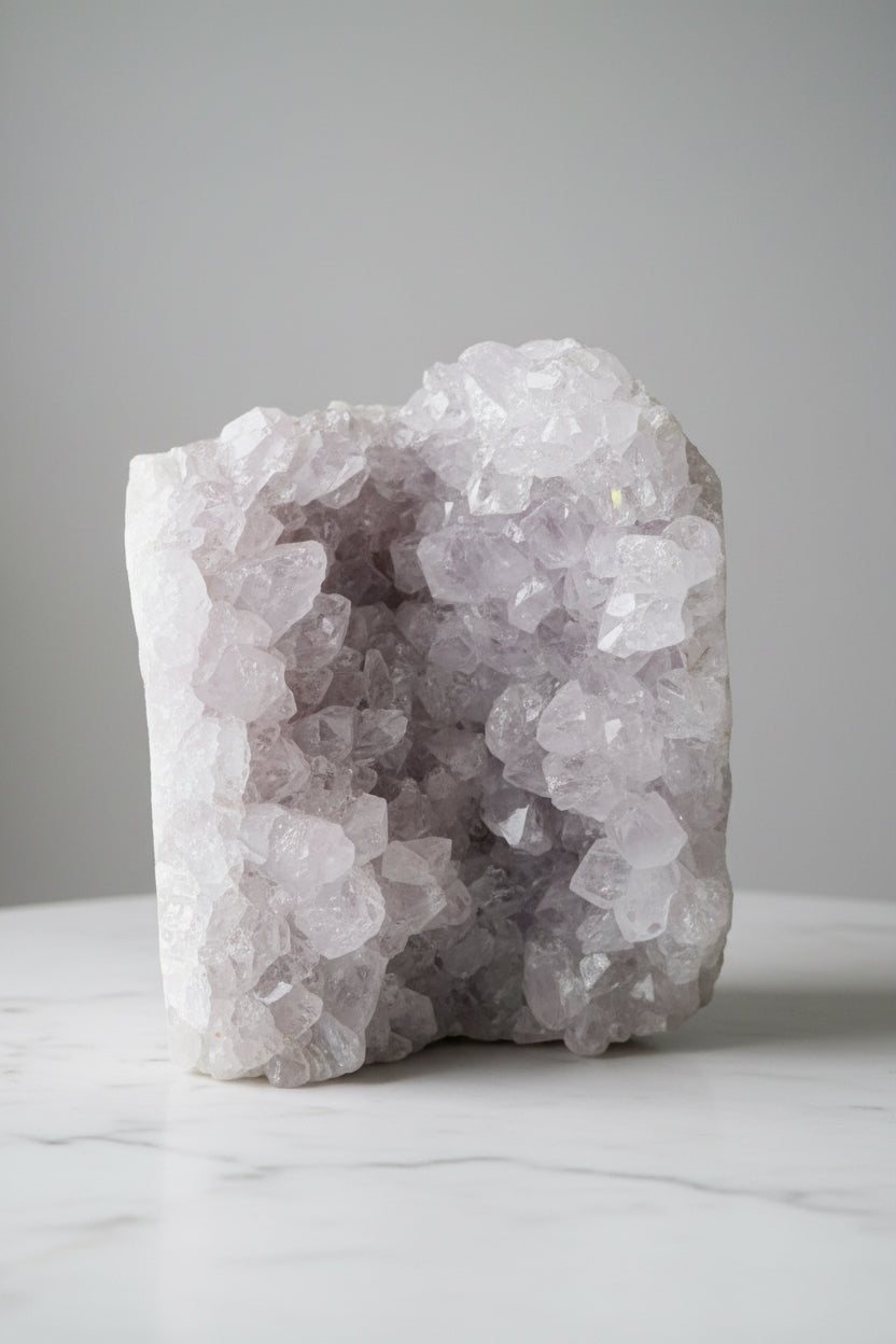 A raw crystal geode specimen with light lavender and white quartz clusters, standing upright on a white marble surface against a neutral gray background.